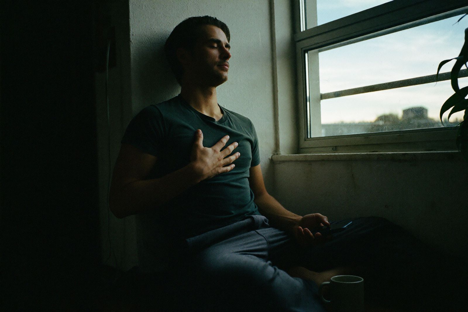 Un hombre junto a la ventana al amanecer, con los ojos cerrados y una mano en el pecho.