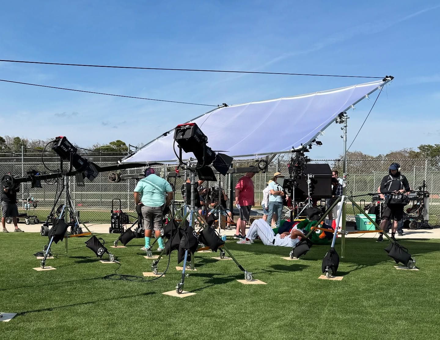 Overhead silk rigged above an exterior set on turf at Fenway South, crew and lights on stands below