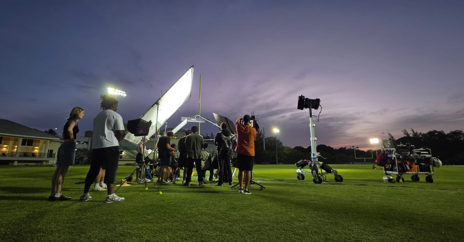Crew on a field at dusk with a lit frame and lights on stands, sunset behind them