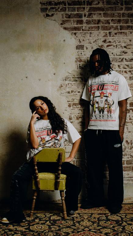 Two models in graphic tees against an exposed brick wall