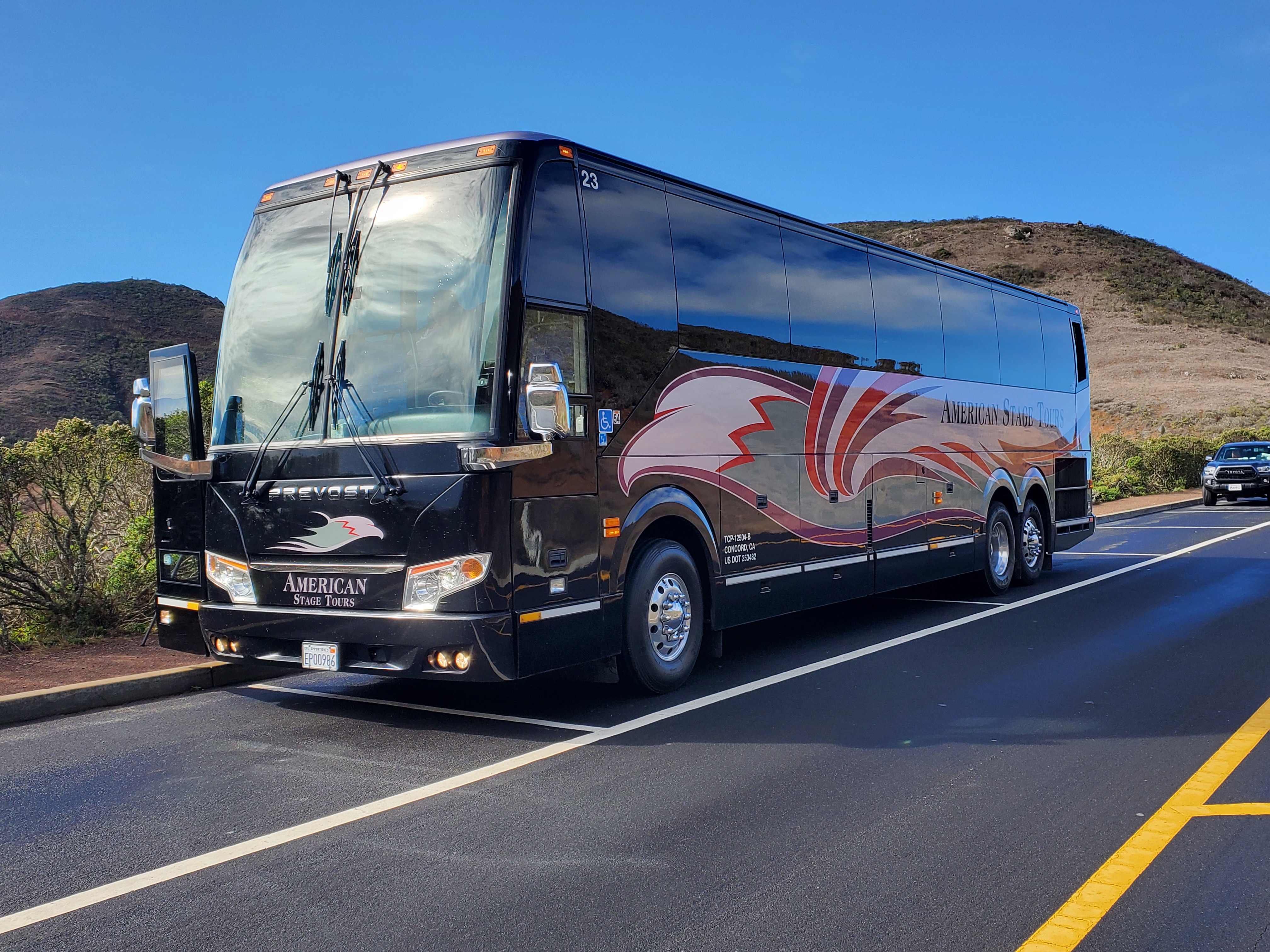 An American Stage Tours coach parked on a coastal road below open hills.