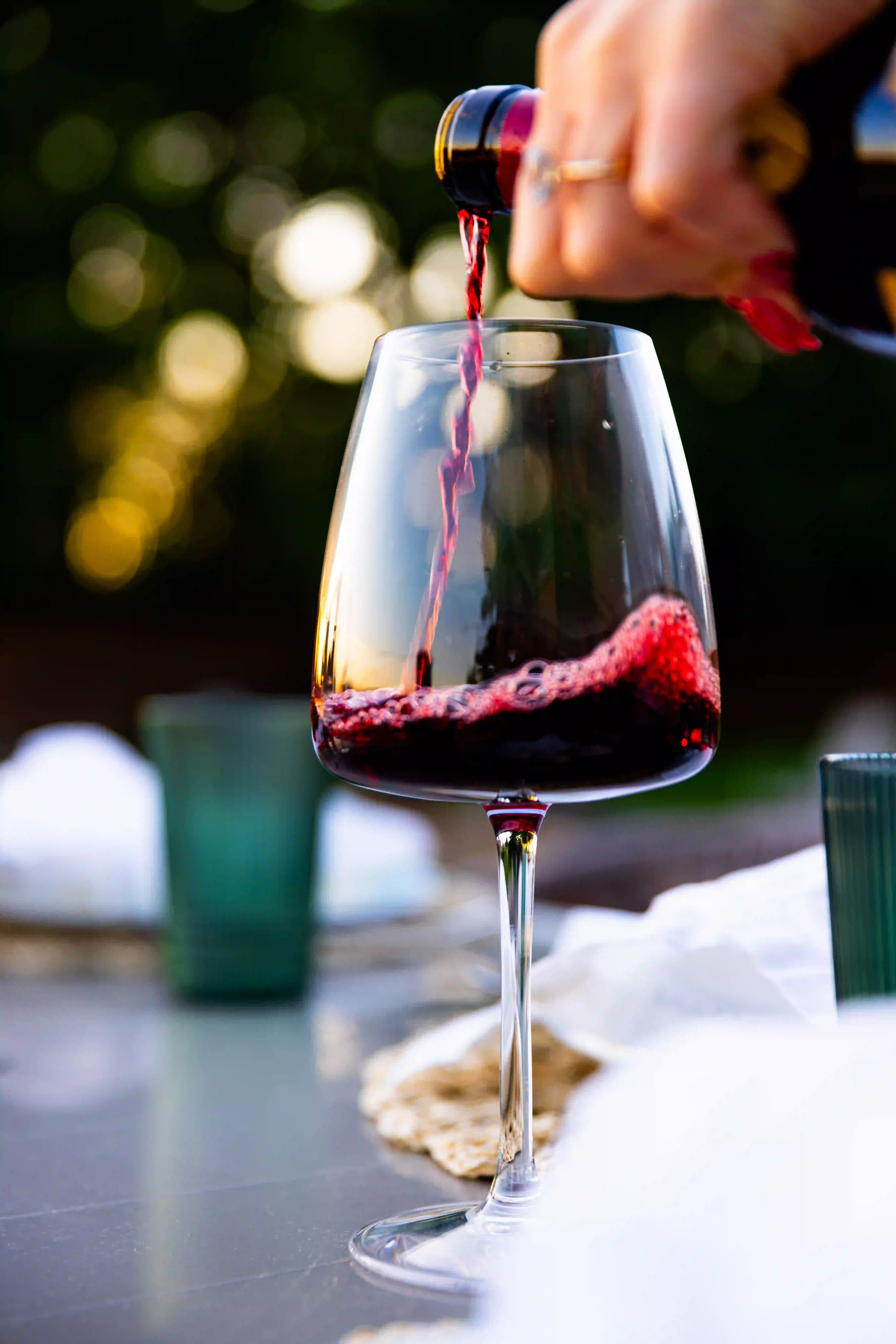 Luxury red wine being poured into a clear stemmed wine glass outdoors at sunset, with soft bokeh lighting and an elegant dining table setting in the background.