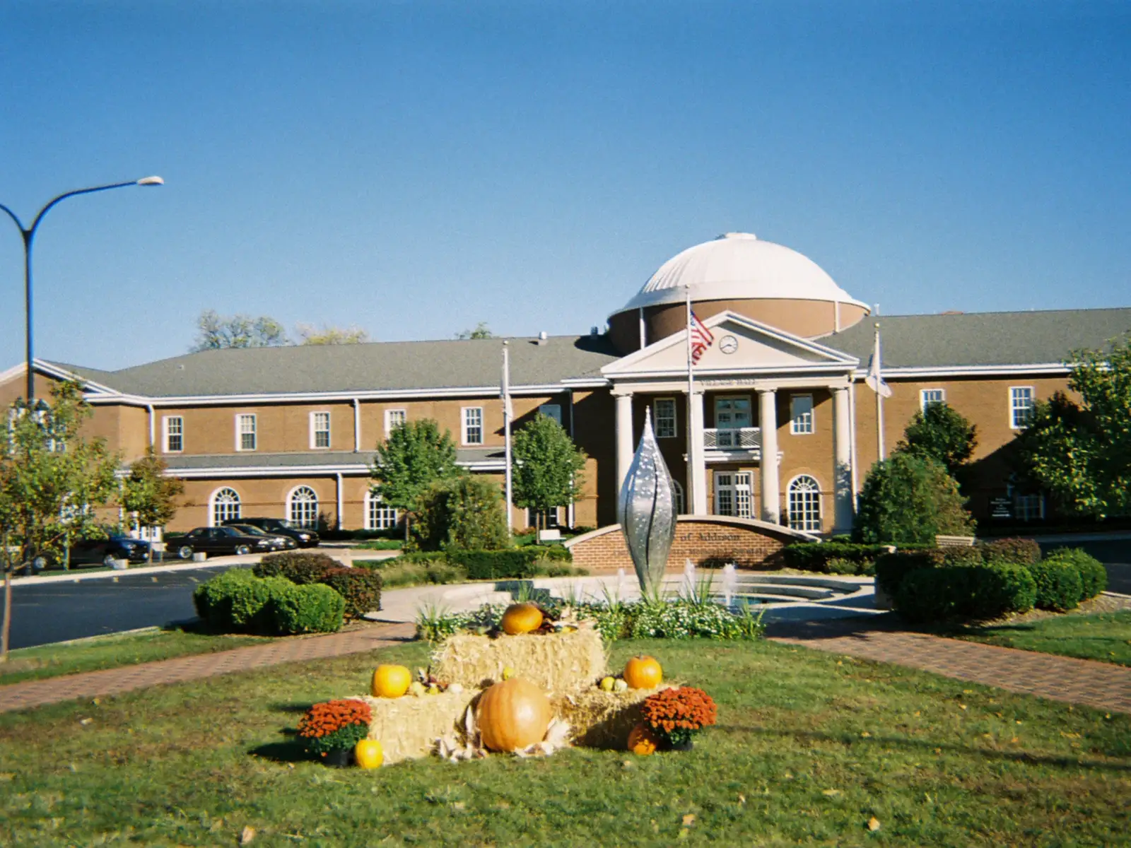 Addison Village Hall, a two-story brick building with a white dome and columned entrance, behind a lawn with a fountain and a fall display of hay bales and pumpkins