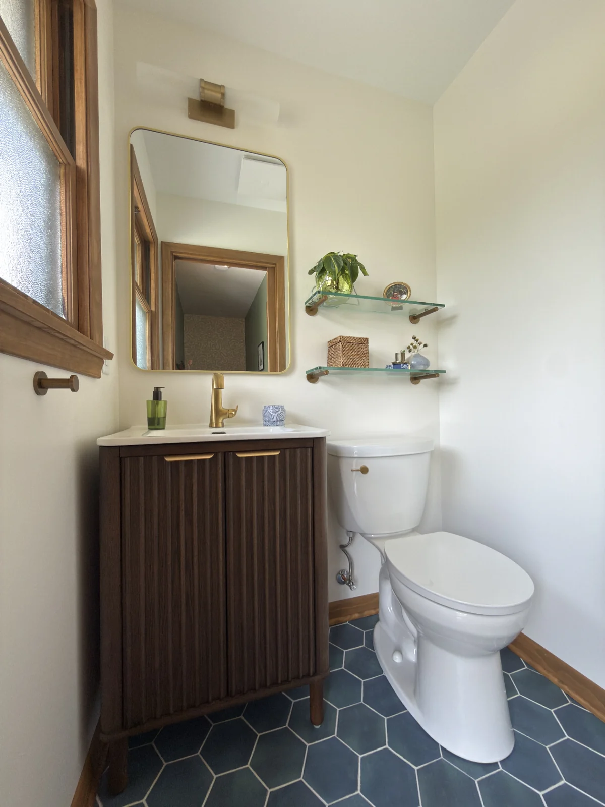 Bathroom after remodel: fluted walnut vanity, brass fixtures, navy hexagon tile floor, glass shelving.
