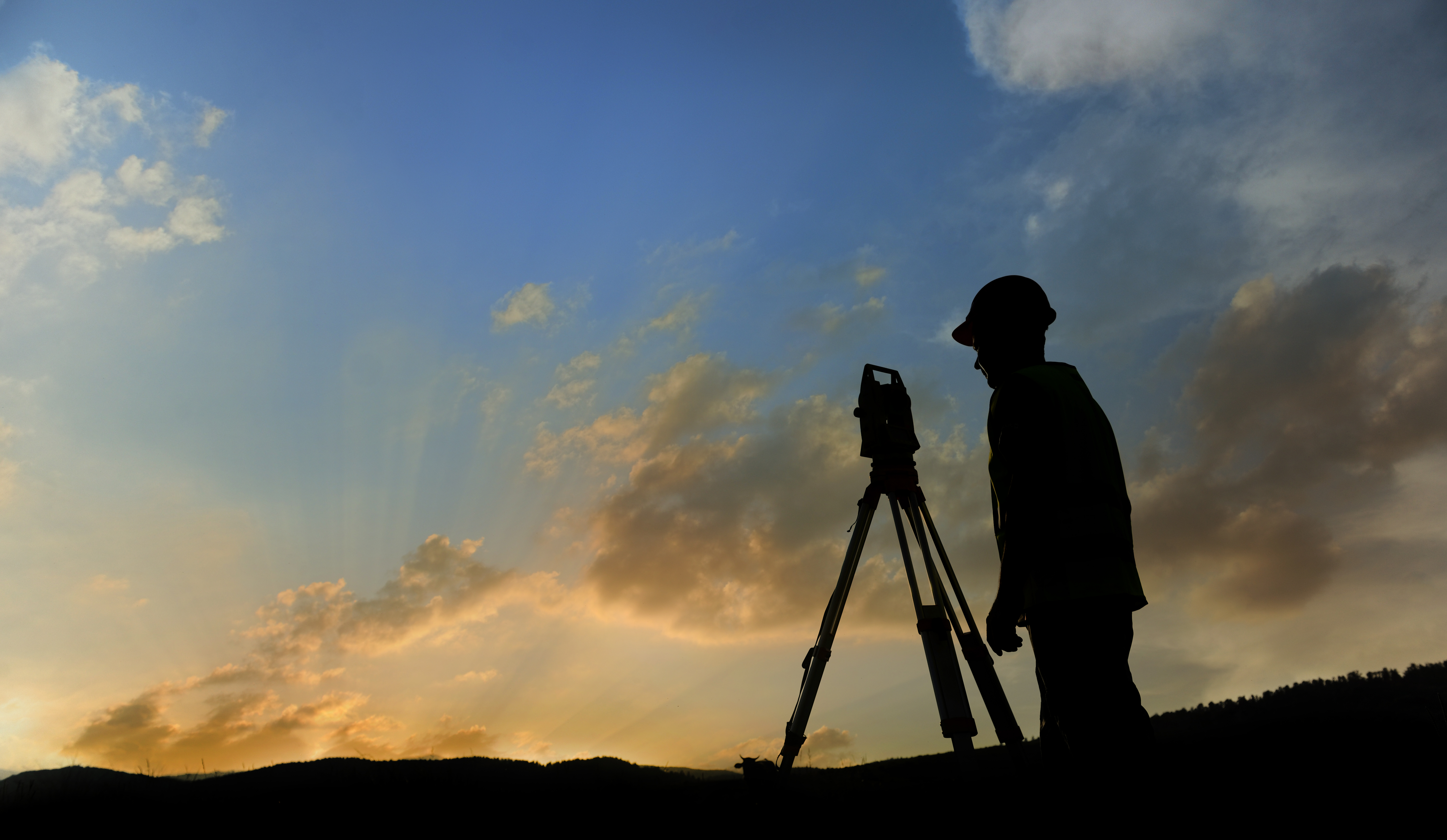 Land surveyor silhouetted beside a tripod-mounted surveying instrument at sunset