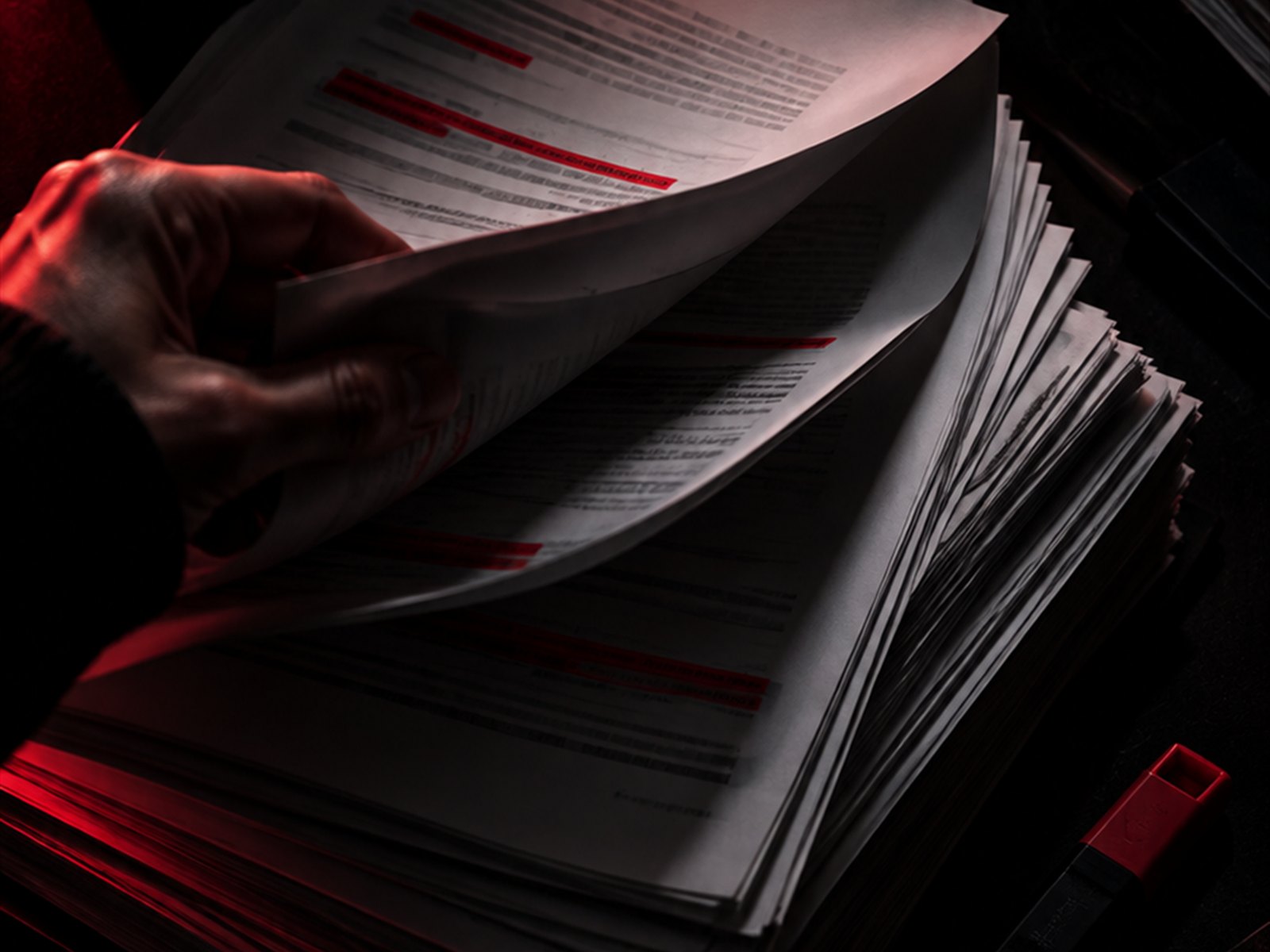 Eine Hand blättert einen hohen Papierstapel durch, einzelne Zeilen rot markiert