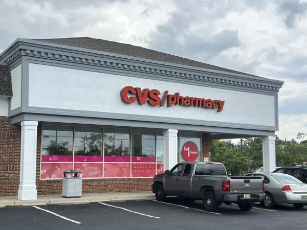 A CVS Pharmacy storefront with a gray truck and silver car parked in front on a cloudy day.