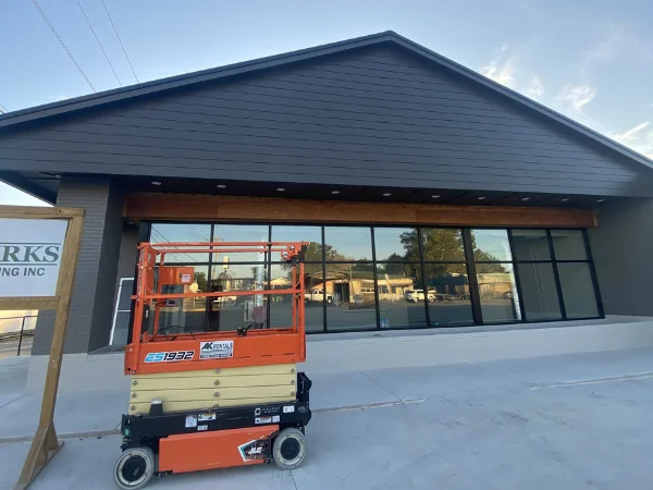 An orange scissor lift sits in front of a newly constructed building with dark siding and large windows.