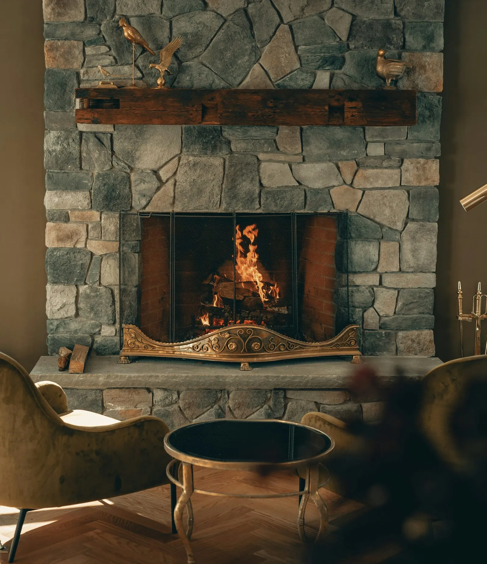 A fire burning in a stone fireplace with a reclaimed wood mantel, a brass fire screen and two velvet armchairs