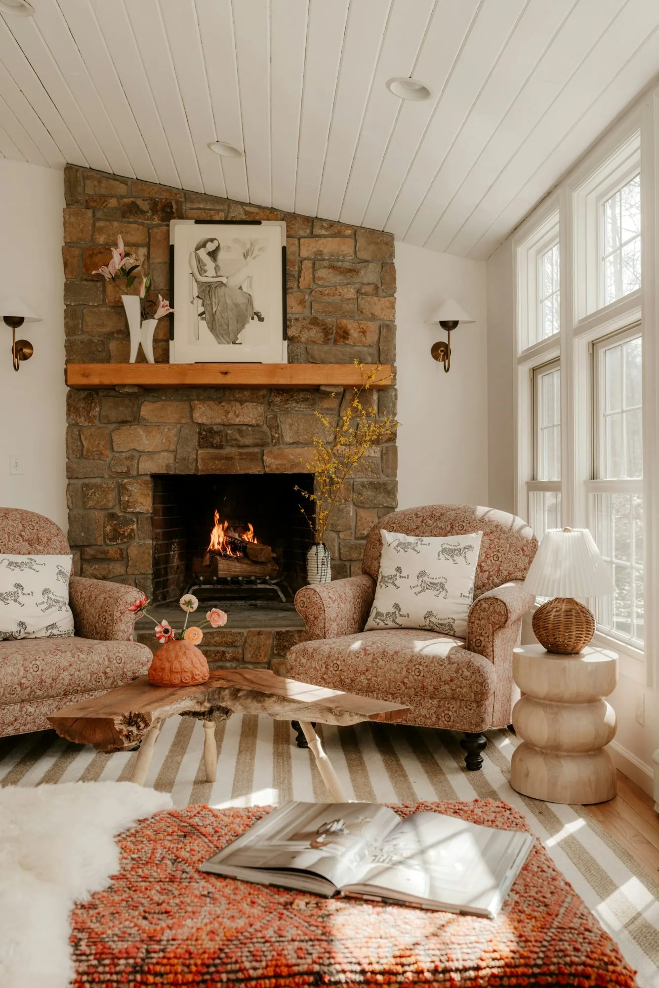 A sunlit sitting room with a stone fireplace, a fire lit, two patterned armchairs, a live-edge coffee table and tall windows