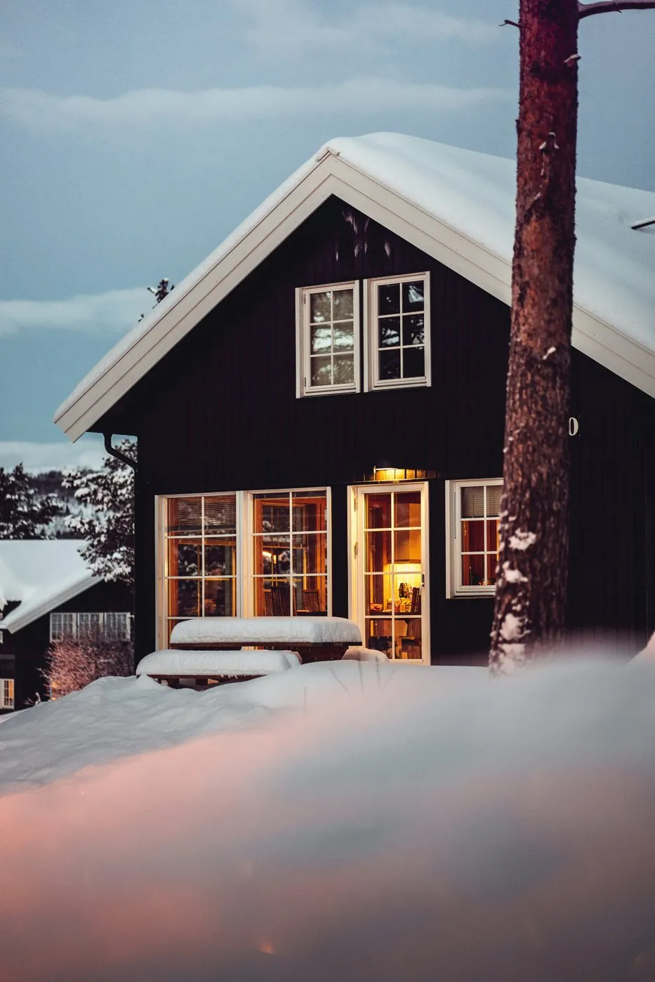 A dark-sided house with warm lit windows above deep snow at dusk