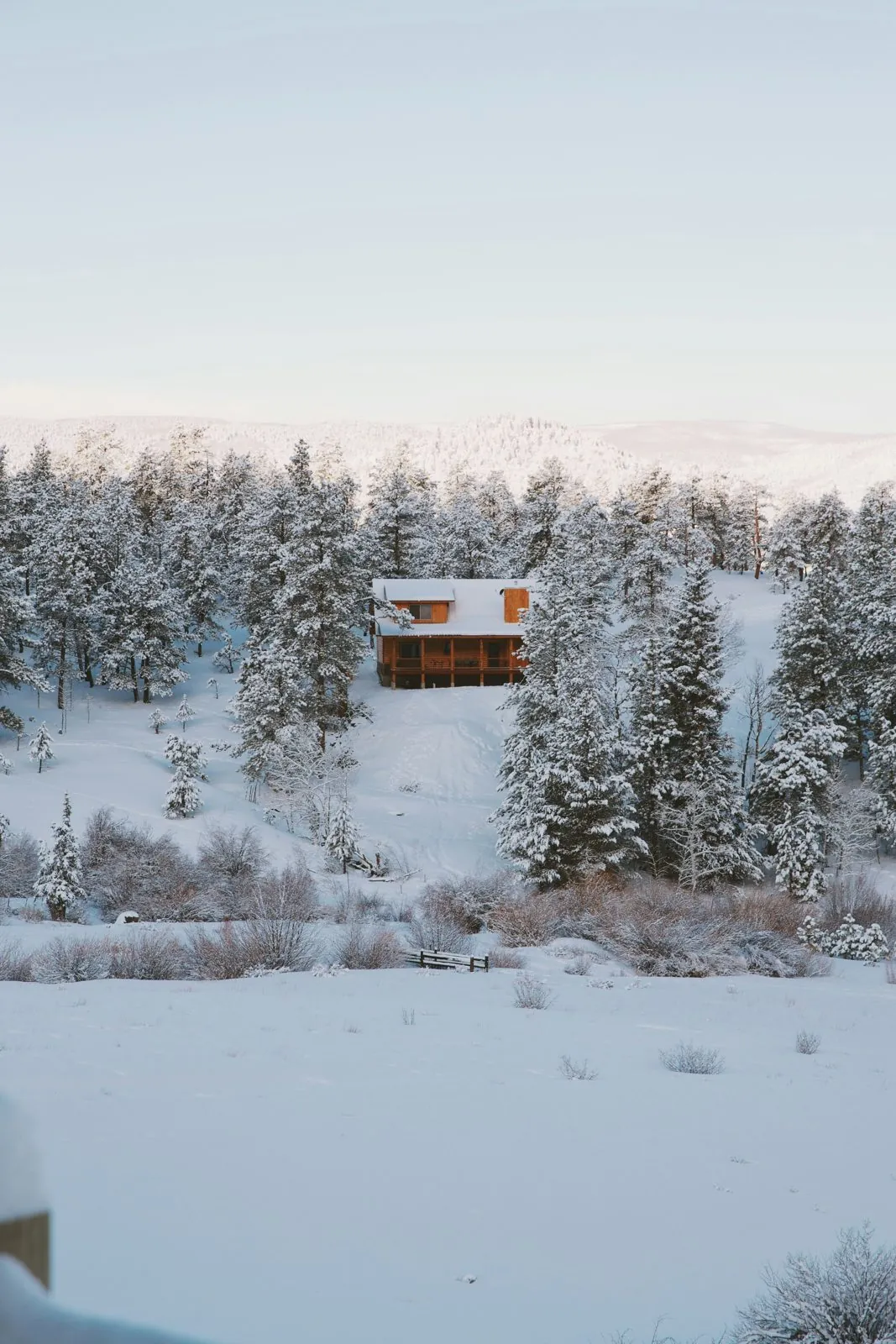 A wooden house on a snowy hillside among snow-covered pines, with soft winter light over the hills