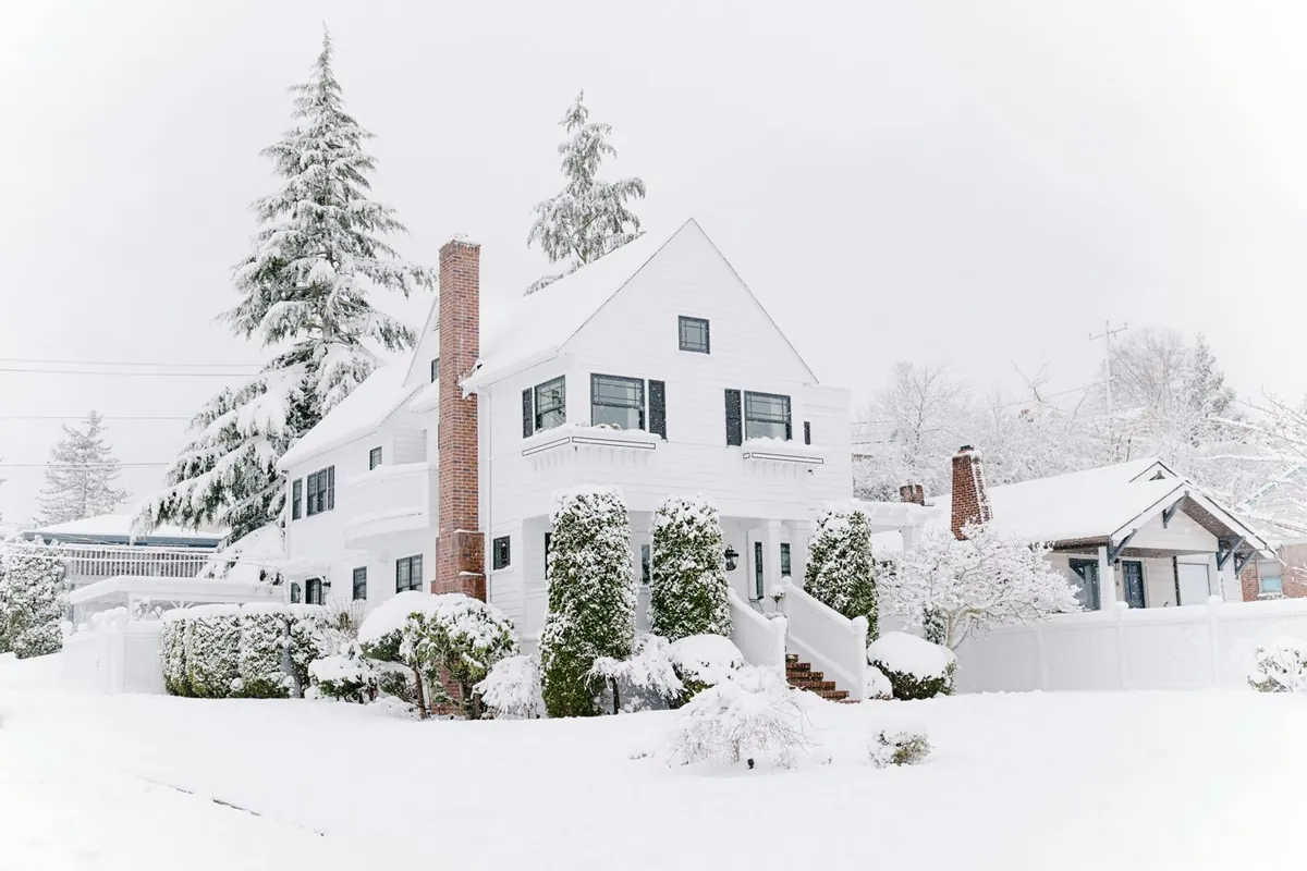 A white farmhouse with black shutters and a brick chimney after a heavy snowfall