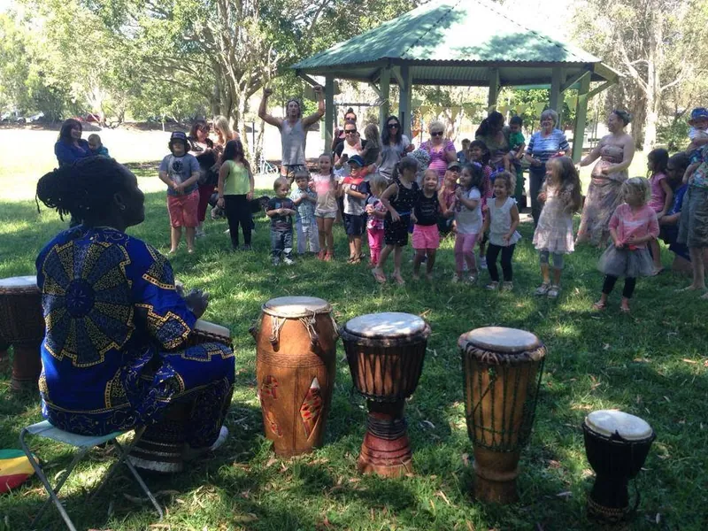 Children and parents seated on the grass with djembe drums lined up at an African Beat Jungle Beats party, where everyone has a turn on the drums