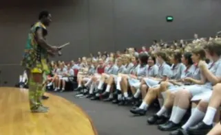 African Beat performer on stage at a high school performance, with rows of students in uniform