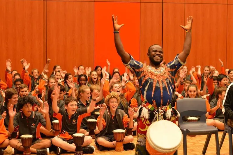 African Beat drummer with arms raised in front of a hall full of students playing drums