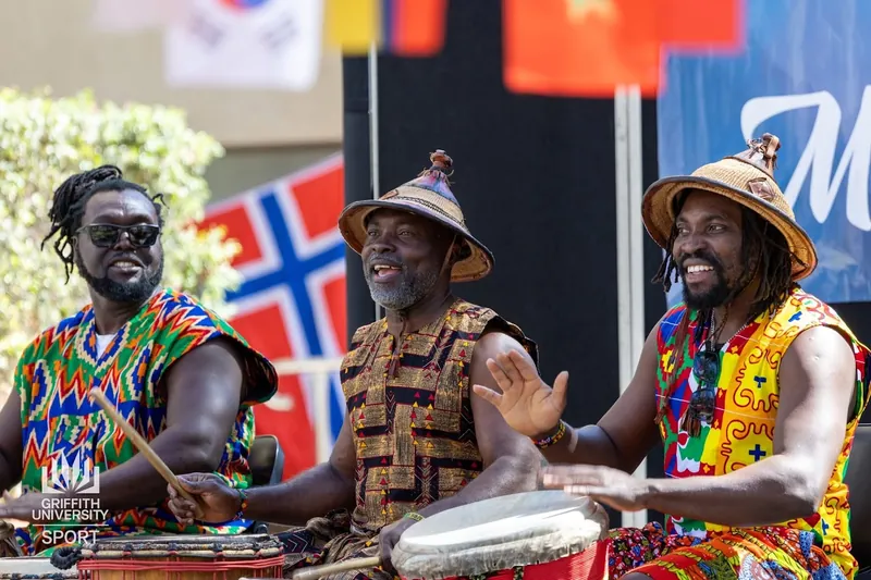Three African Beat drummers in kente cloth performing in front of national flags