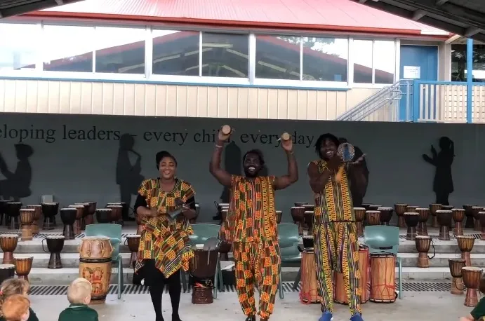 African Beat performers teaching primary school children about the instruments, with rows of drums outside the school