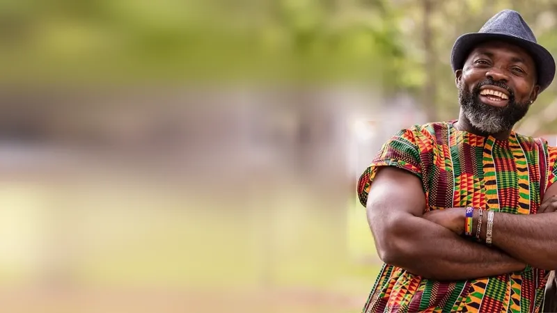 Master Tuza smiling in kente shirt and hat against an outdoor green backdrop