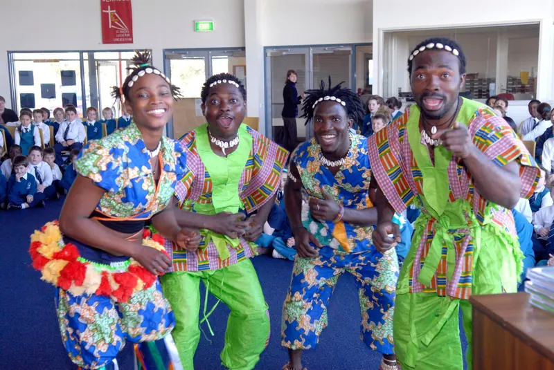 Four smiling African Beat performers in bright traditional African prints and cowrie-shell headbands dancing in a school hall in front of seated students in uniform
