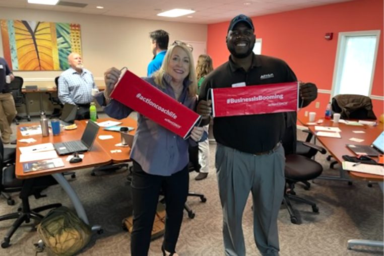 Master Coach Mary Ann Hauser celebrating with a business owner holding #actioncoachlife and #BusinessIsBooming banners