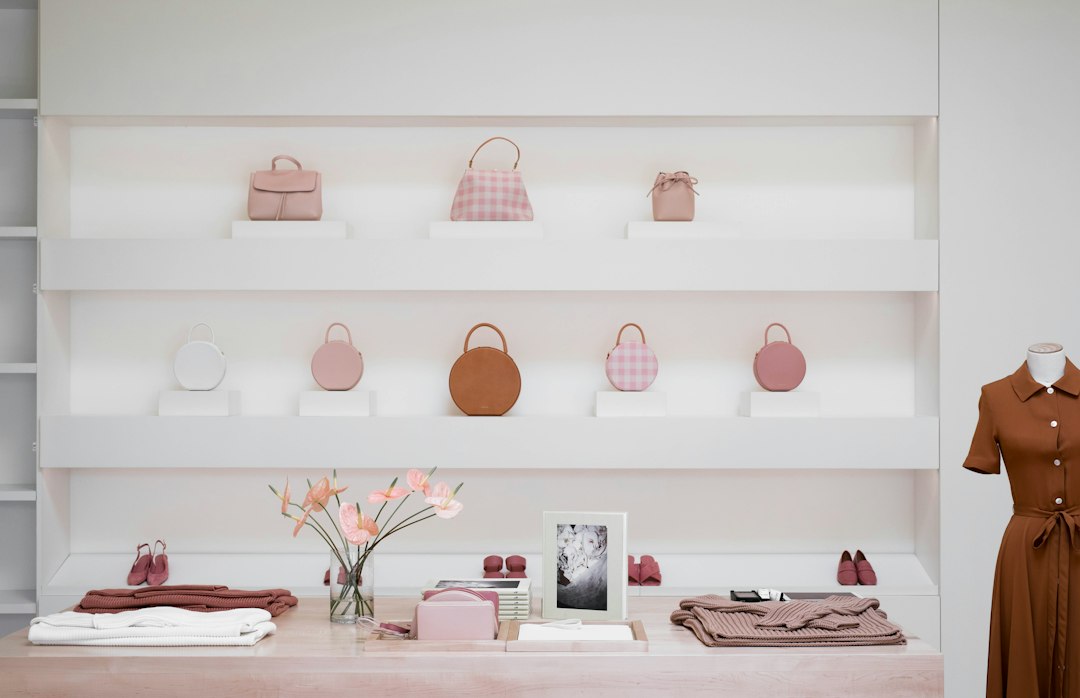 Bright white retail shelving displaying pastel handbags and shoes above a wood table with folded clothing and flowers.
