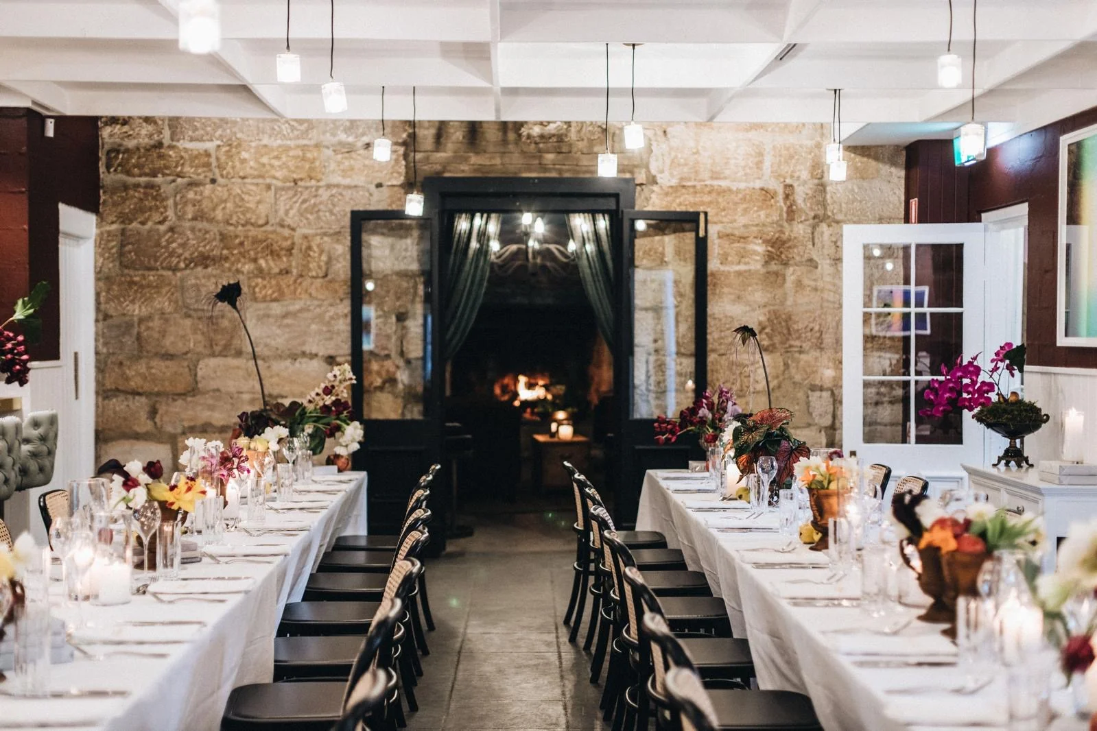 Long dining tables with white tablecloths and flowers beside a stone fireplace