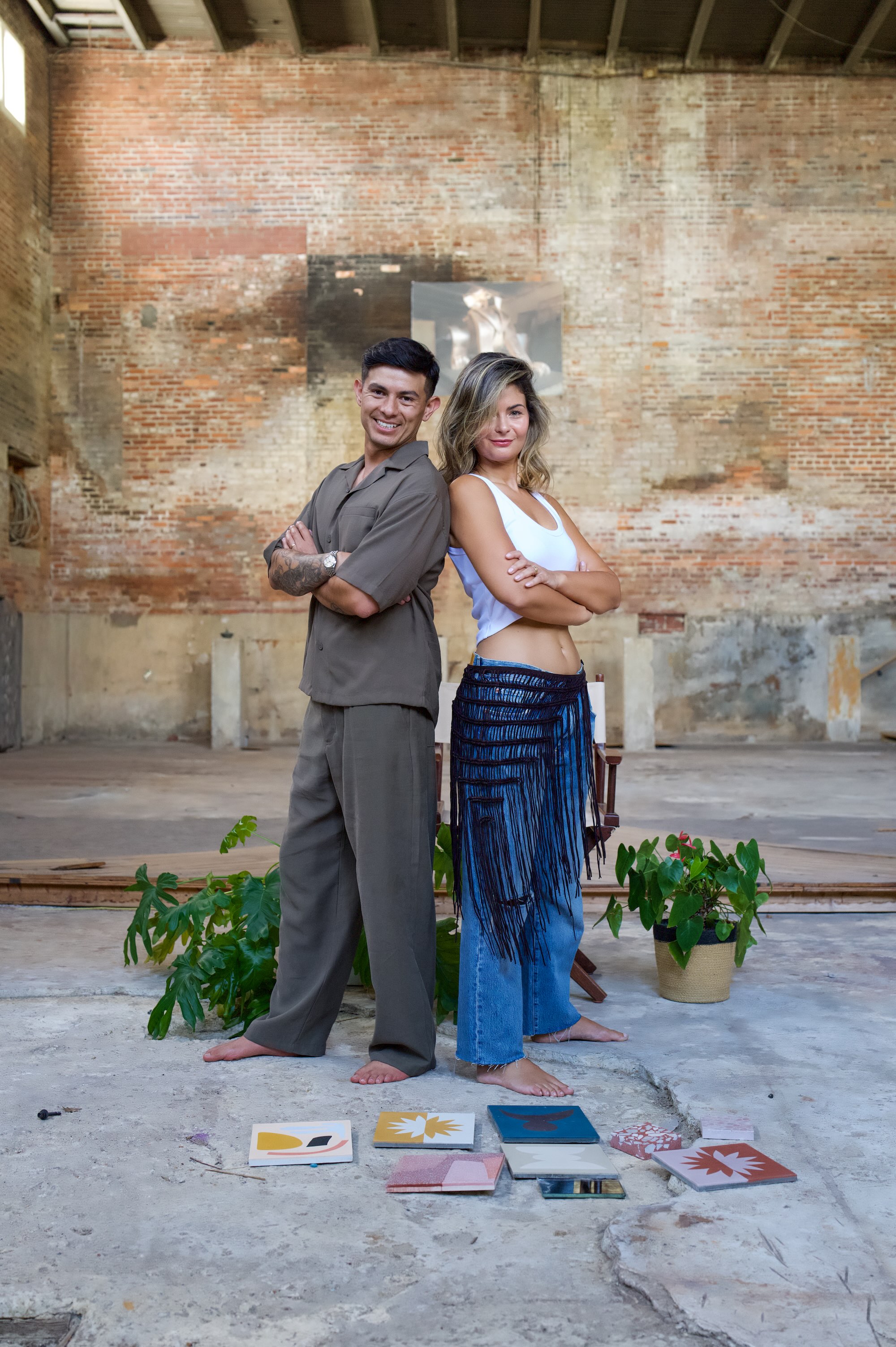 Ana Clara Sabena and Javier Suarez, founders of A Couple & A Dream — back-to-back with arms crossed in a brick warehouse setting.