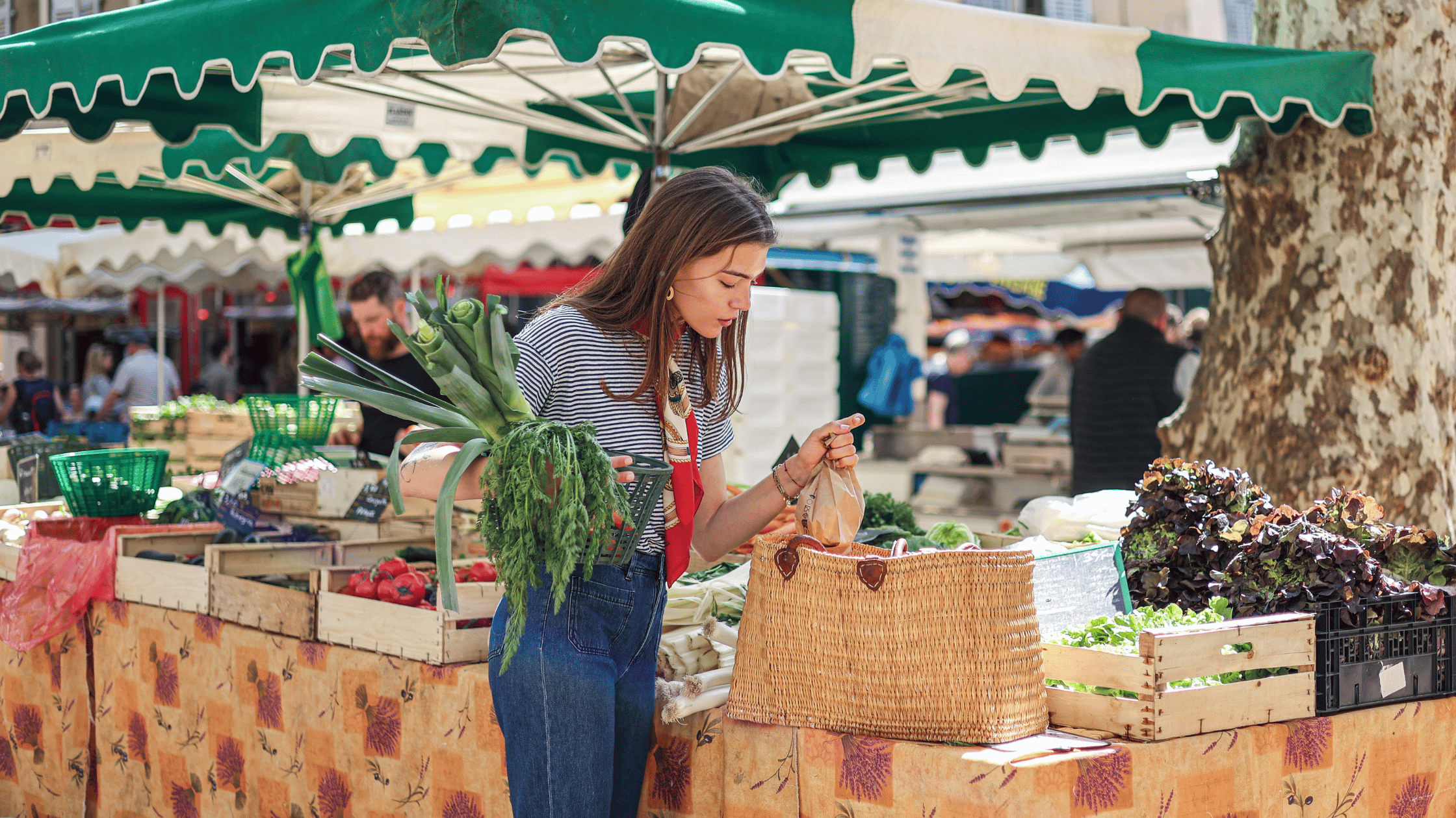 A person shops for fresh produce at an outdoor market, holding a bunch of leeks and a paper bag over a woven basket.