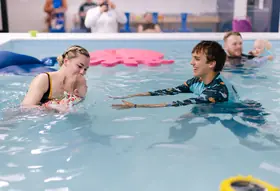 Instructor kisses the top of a baby's head while a parent films in the background