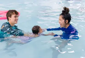Instructor reaches out with open hands as a colleague floats a baby gently by