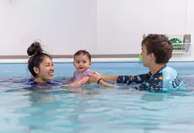 Two instructors laughing together as they support a dark-haired swimmer