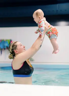 Coach lifts a baby overhead in the pool, beaming up at the child