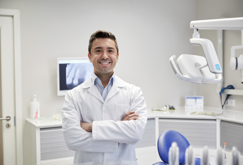 Alt text: Smiling dentist standing with arms crossed in a clean dental clinic, with dental chair and equipment visible in the background, representing a professional healthcare provider ready to assist patients