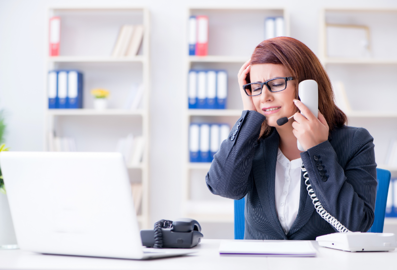Alt text: Frustrated businesswoman wearing a headset and holding a phone while working at a desk with a laptop, appearing stressed by incoming calls and overwhelmed with customer inquiries