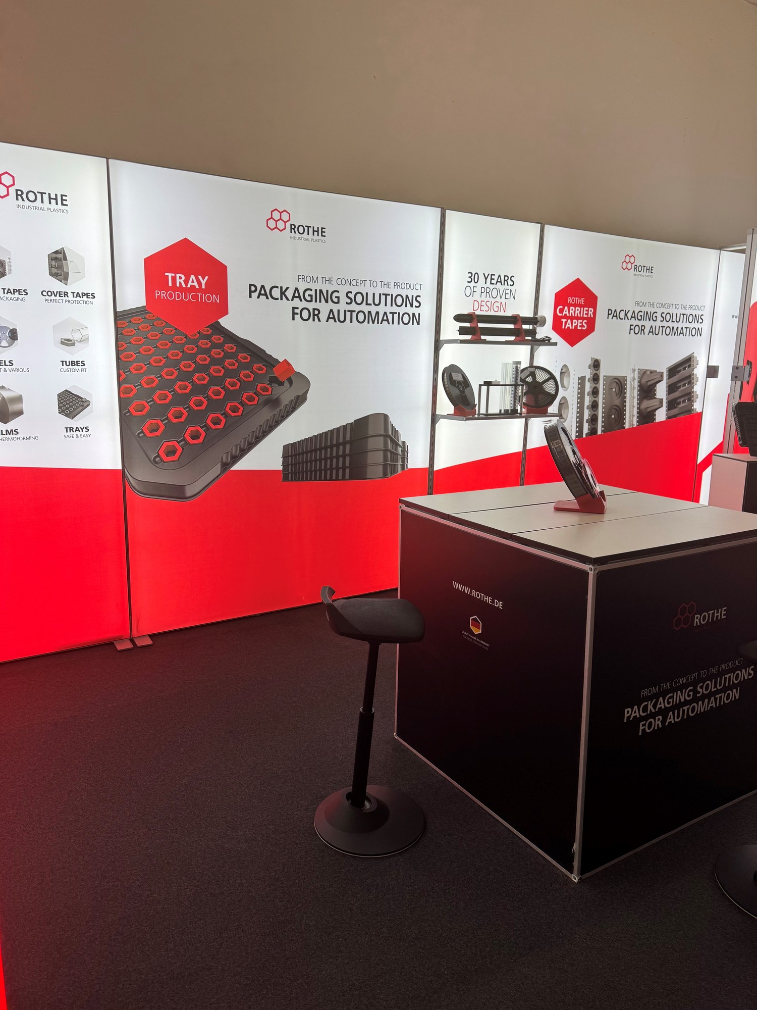Schwarzer Würfeltisch mit Hocker vor der Wand „Packaging Solutions for Automation“