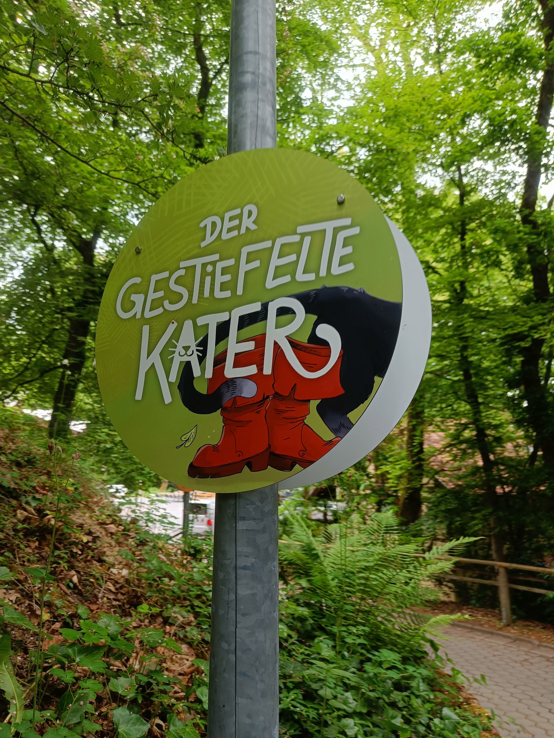 Rundes grünes Schild mit Schriftzug und roten Stiefeln am Waldweg