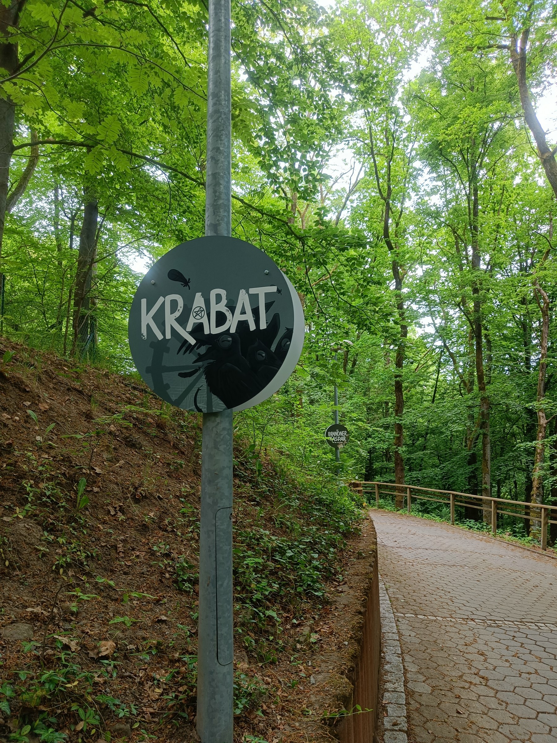Rundes dunkles Schild mit Krabat-Schriftzug an einem Pfosten am Waldweg