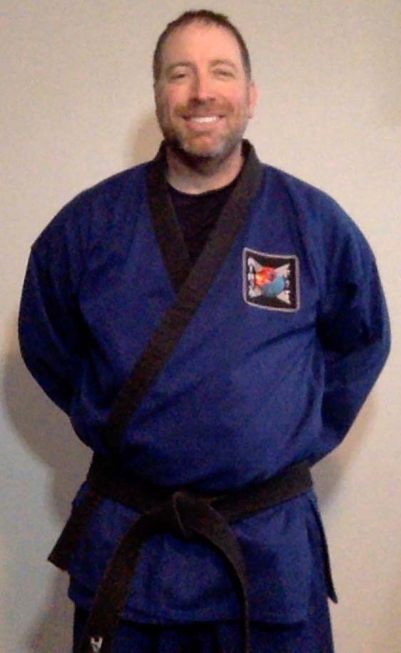 martial arts instructor in blue uniform and black belt smiling while standing with hands behind his back