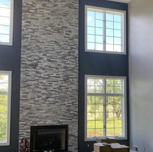 Modern living room interior featuring a tall, gray stone fireplace surrounded by large windows with white frames, allowing natural light to fill the space.