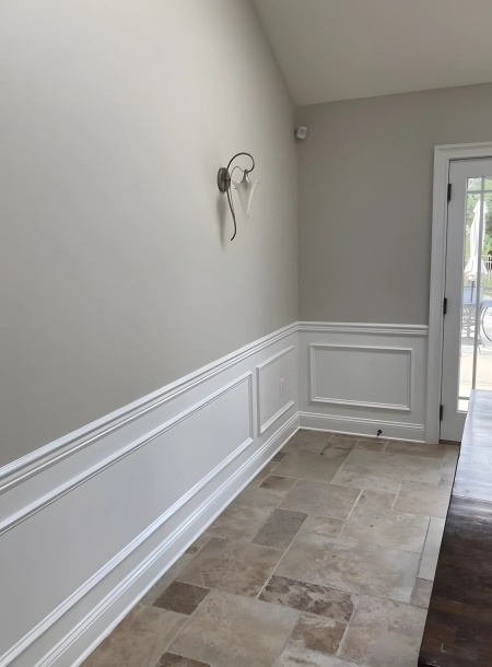 Light gray wall with white wainscoting and molding, featuring a modern wall sconce and beige tiled flooring in a well-lit hallway with a door leading outside.