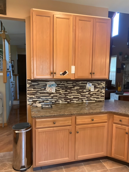 Light wood kitchen cabinets with a granite countertop, featuring a modern mosaic tile backsplash in neutral tones. A stainless steel trash can is placed next to the counter, and the kitchen has an open layout leading into the living area.