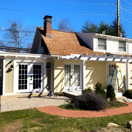 Exterior view of a light yellow house with a white pergola over the patio, featuring French doors, a brick walkway, and a landscaped garden. A ladder and tools are set up for ongoing work, and the roof has reddish-brown shingles.