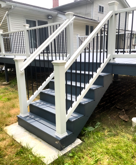 Newly constructed outdoor deck stairs with white posts, black metal railings, and gray steps, leading to a raised deck attached to a white house. The yard surrounding the deck features grass and a concrete landing at the base of the stairs.