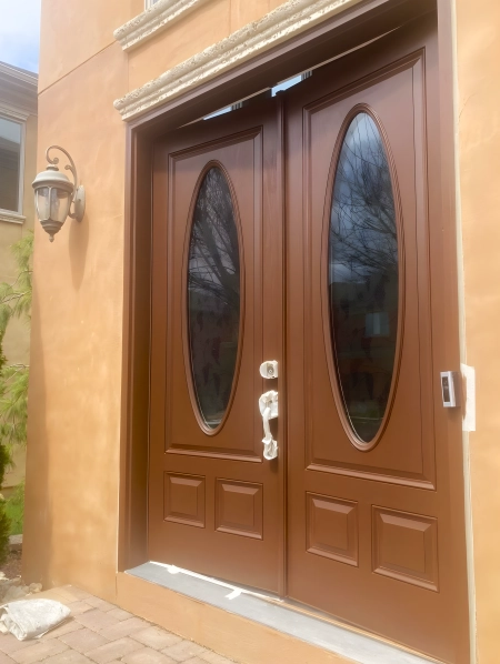 Double brown wooden front doors with oval glass panels, framed by a beige stucco exterior wall. The entryway is adorned with a decorative wall-mounted light fixture and stone accents above the doors.