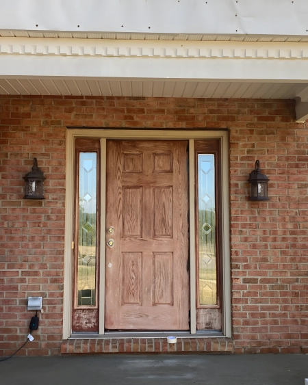 Front entrance of a brick house featuring a weathered wooden door with glass side panels, flanked by two outdoor wall lanterns.