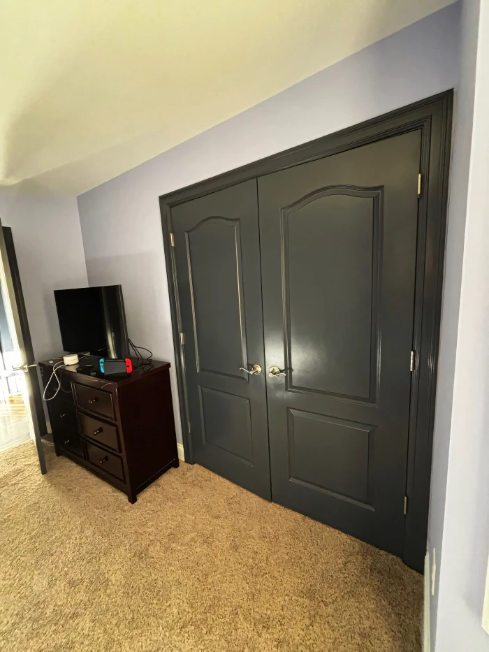 Dark gray double doors with a smooth finish in a freshly painted bedroom featuring light purple walls and beige carpet flooring.