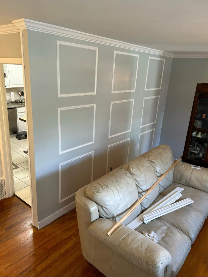 Light blue interior wall with decorative white rectangular trim accents, freshly painted, next to a beige leather sofa and wooden floor, with trim pieces placed on the sofa. A kitchen doorway is visible on the left.