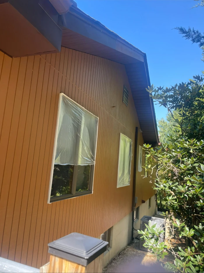 Freshly painted orange-brown exterior wall of a house with protected windows covered in plastic sheeting, surrounded by green trees under a clear blue sky.