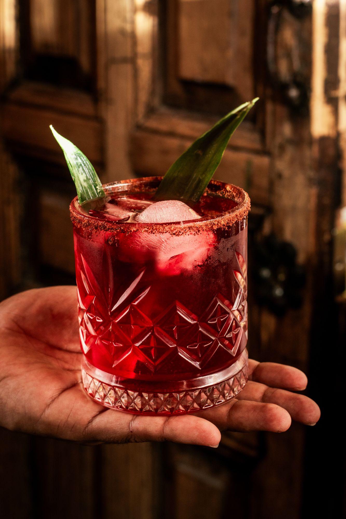 A red house cocktail with a chile rim and a pineapple-leaf garnish, held in front of an old wooden door